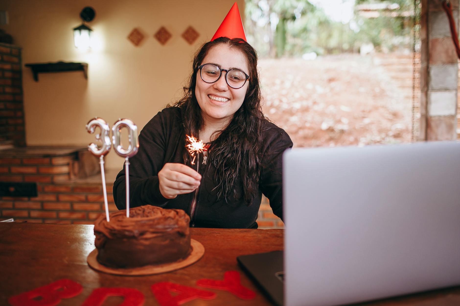 Woman celebrating 30th birthday virtually with cake and sparkler in a cozy indoor setting.