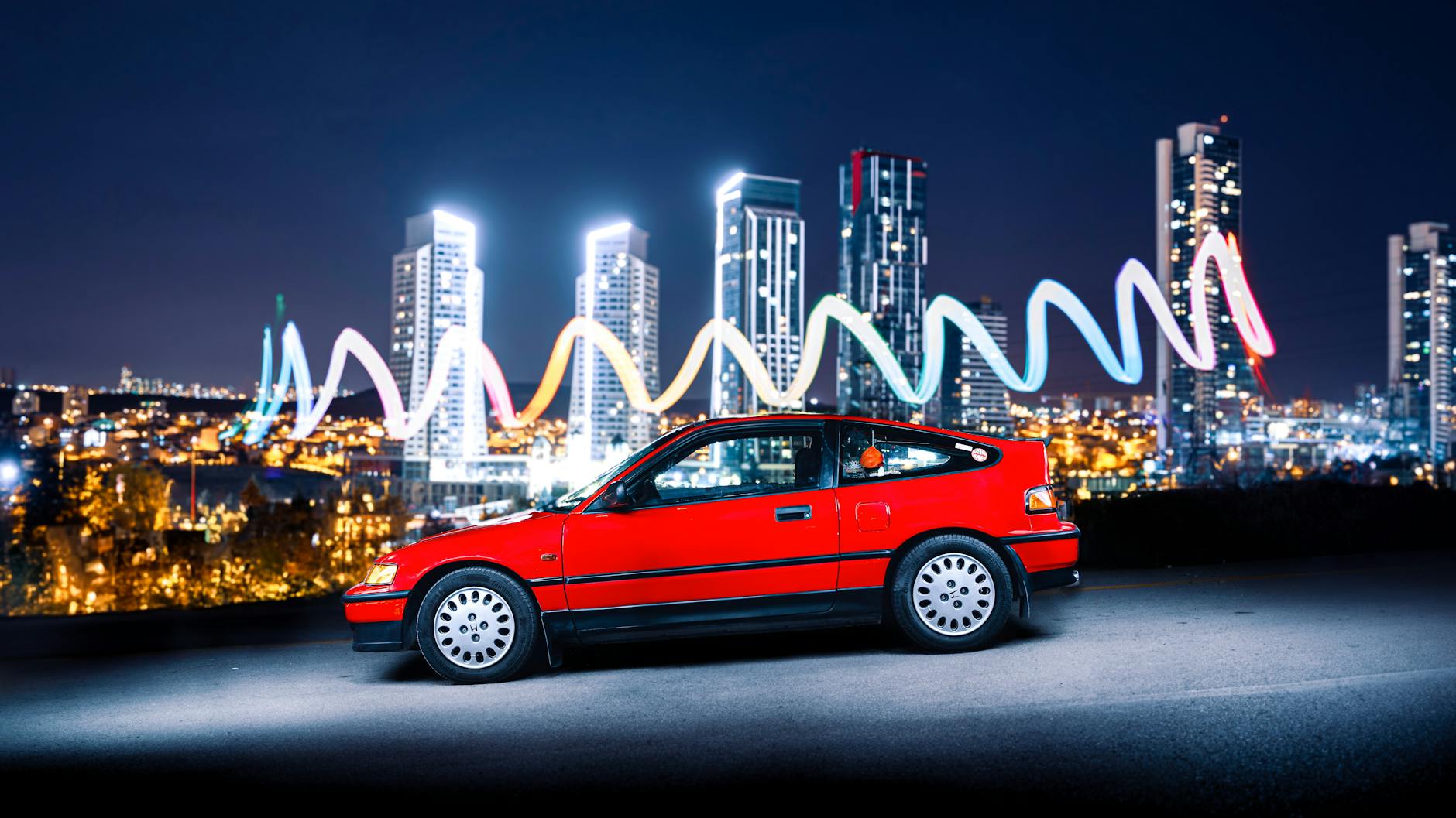 A vibrant red Honda CRX car with Ankara's night skyline as a backdrop, showcasing urban modernity.