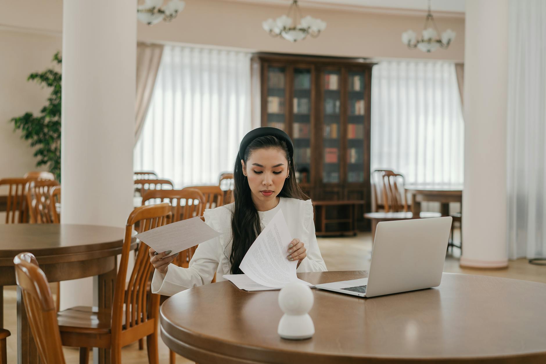 Asian woman using laptop for online learning in a modern library setting, surrounded by books and wooden furniture.