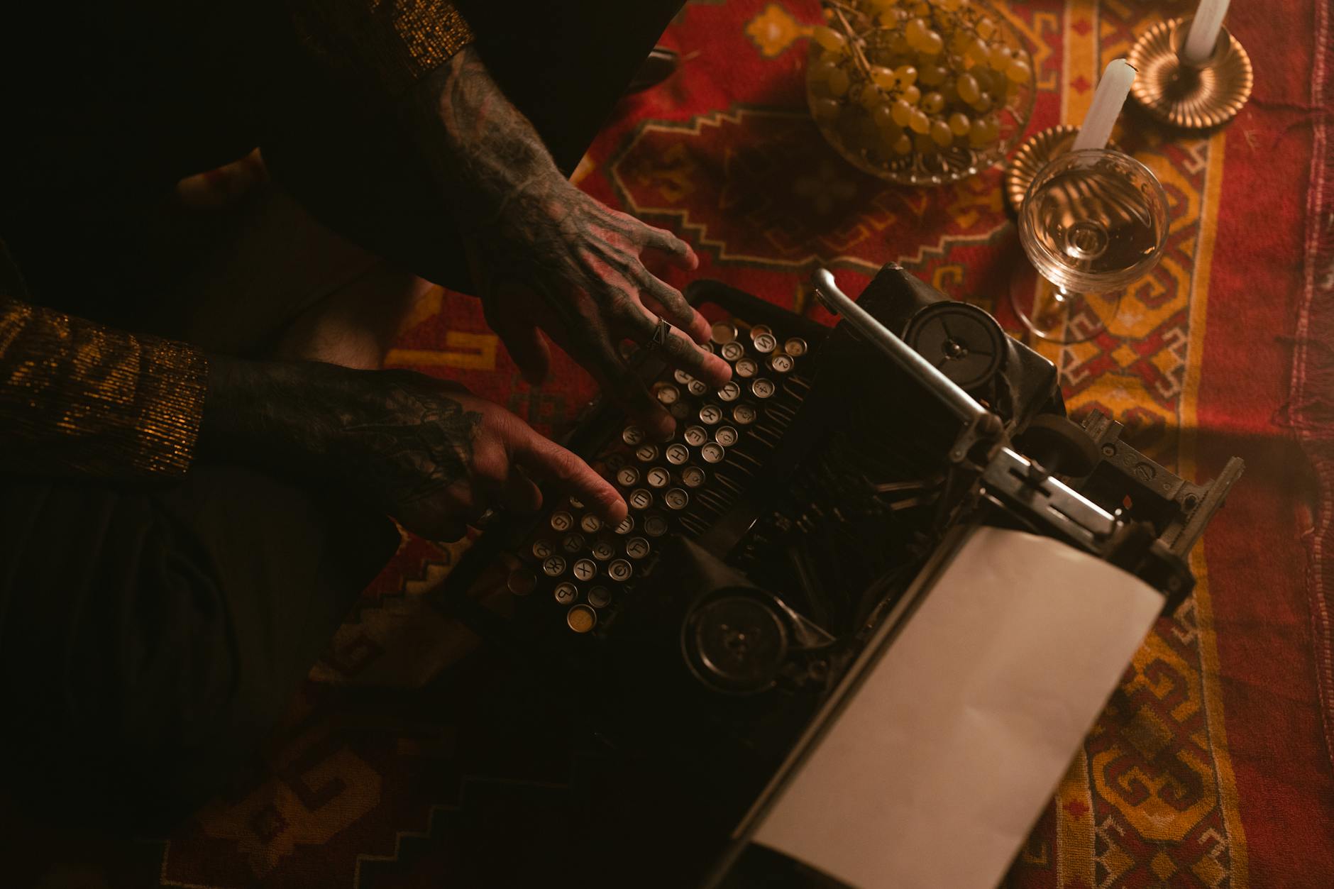 Tattooed hands typing on a vintage typewriter with candles and grapes in a warm setting.