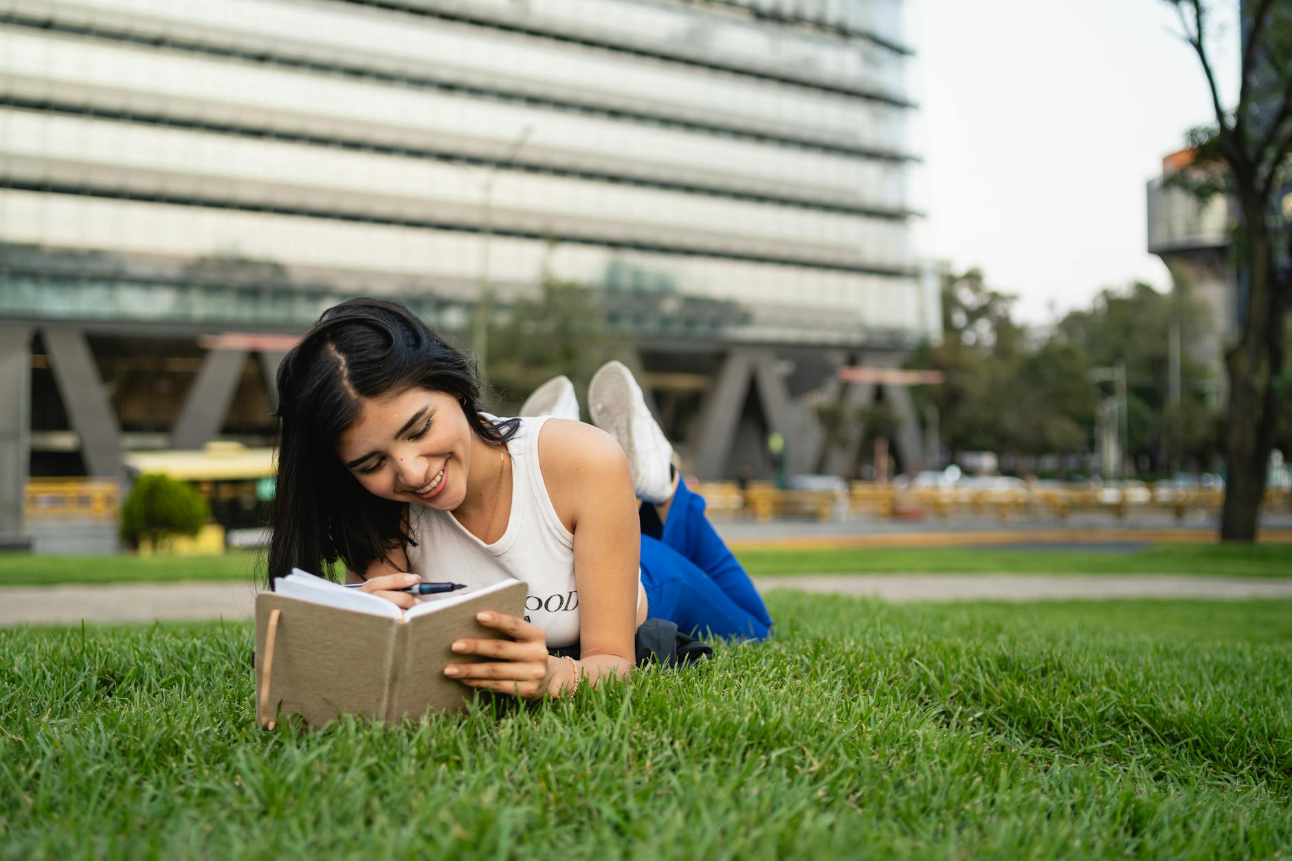 Smiling woman lying on grass reading a notebook in a city park on a sunny day.