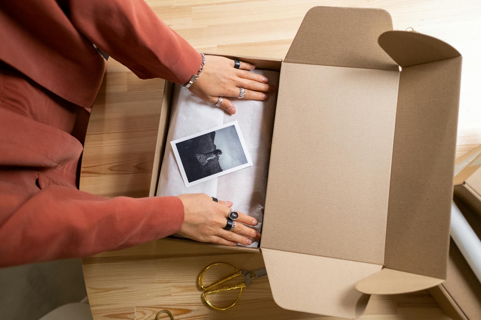 Hands arranging items in a cardboard box with a photo and scissors nearby.