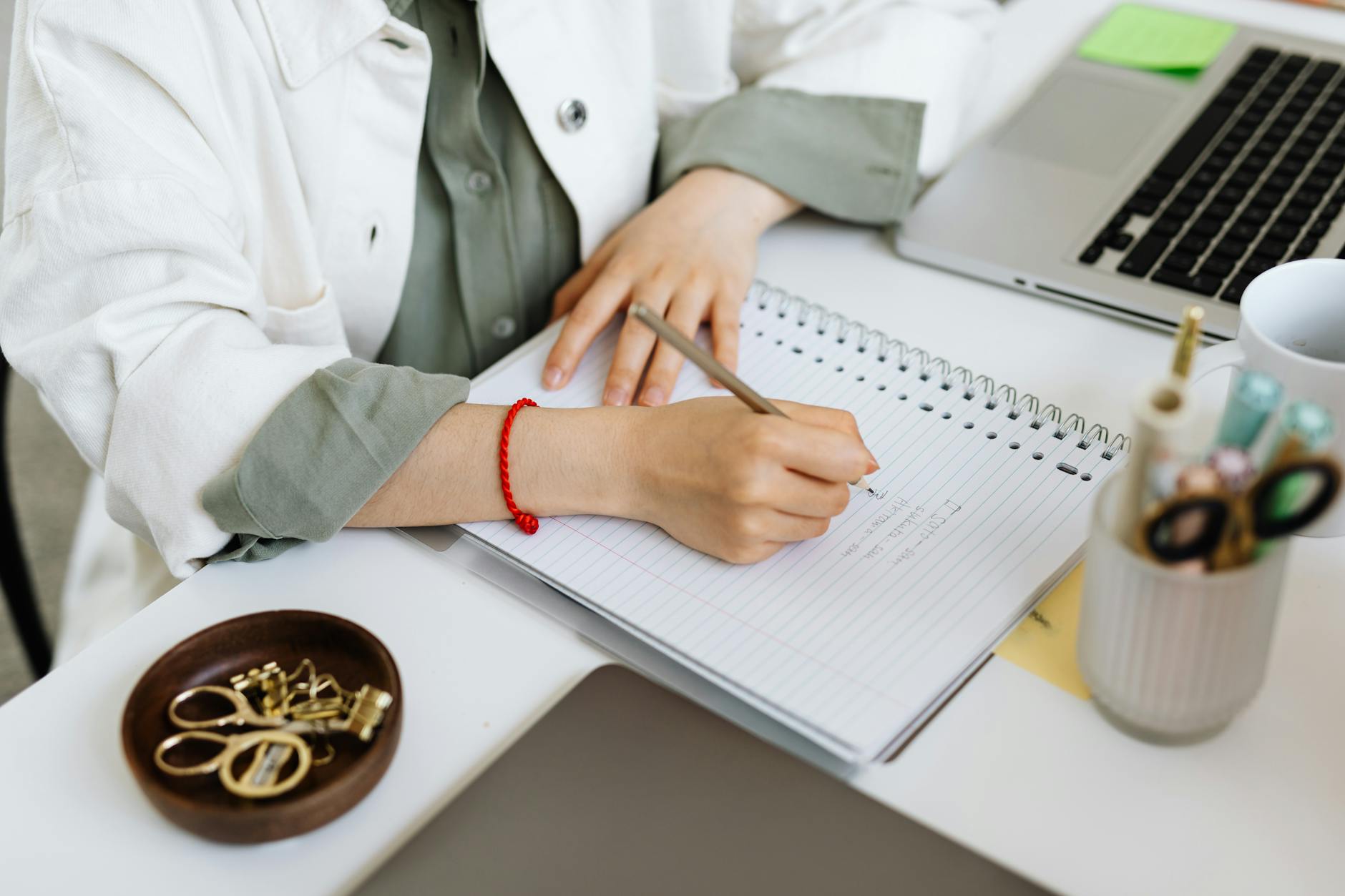 A person writes in a notebook at a desk with office supplies and a laptop, creating a cozy workspace.
