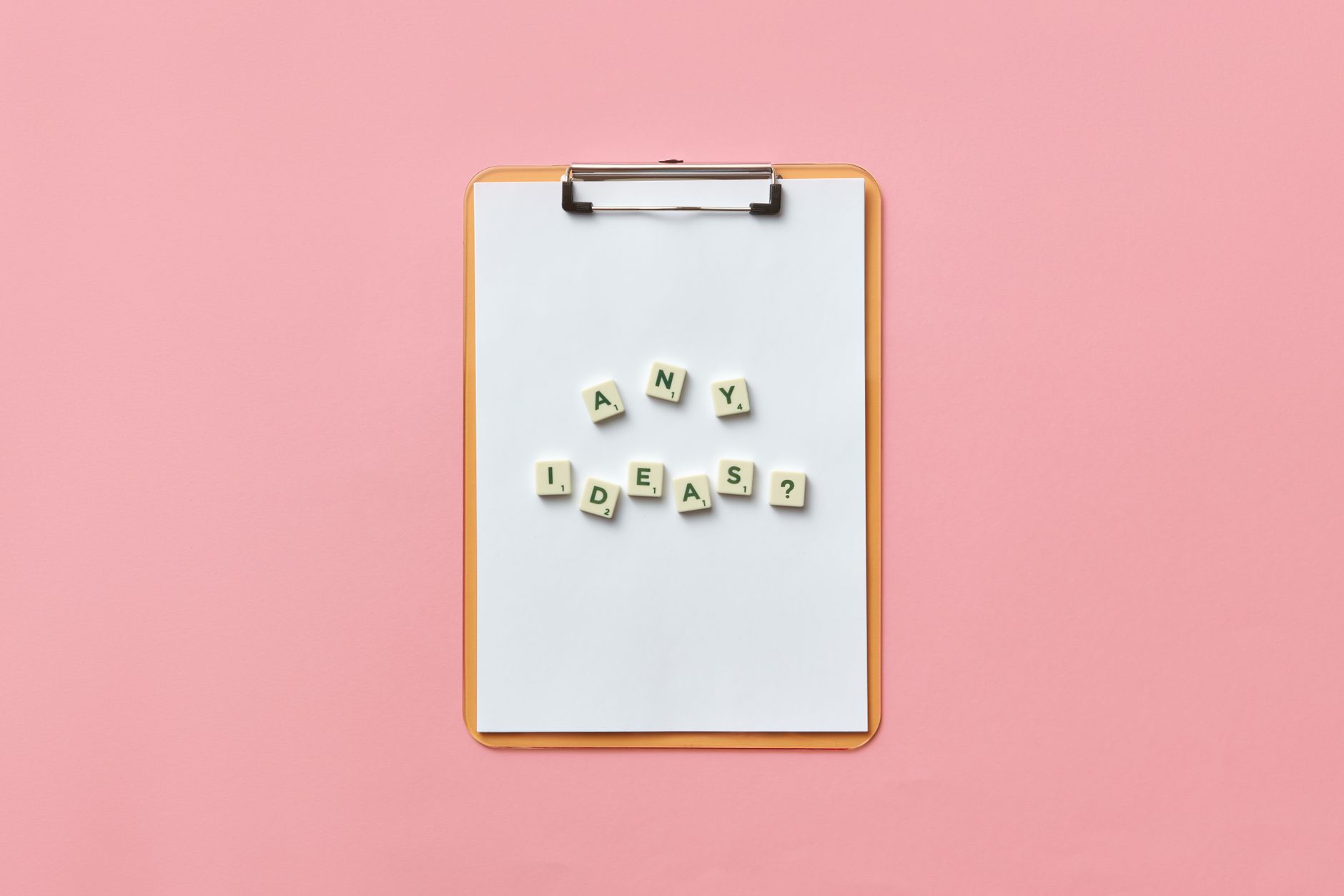 Creative conceptual image of a clipboard with letters spelling 'Any Ideas?' using Scrabble tiles on a pink background.